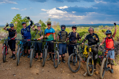 A class of mountain bikers poses for a group picture with a the cloudy sky in the background.