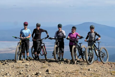 A group of lady mountain bikers pose for a photo on top of a mountain.